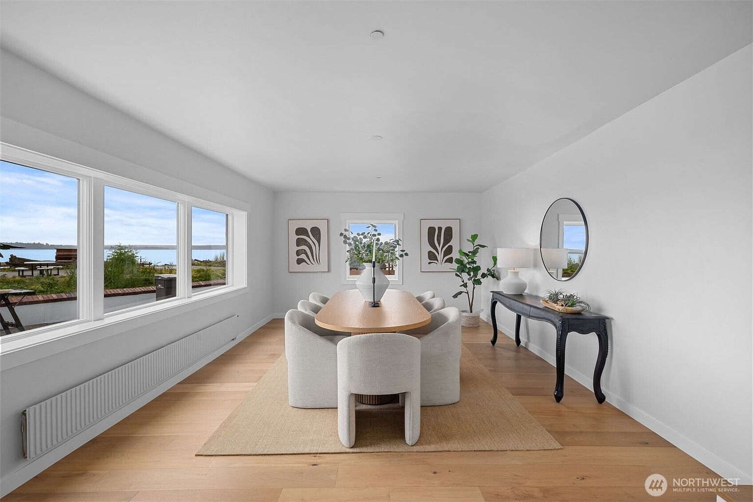 This bright and airy dining room features a minimalist aesthetic with light wood flooring and a large, oval-shaped dining table surrounded by six upholstered chairs. A console table sits against the right wall beneath a round mirror, while two framed botanical prints flank a central window that overlooks a scenic waterfront view. The space is anchored by a large, neutral-toned area rug, creating a serene and inviting atmosphere for formal or casual dining.
