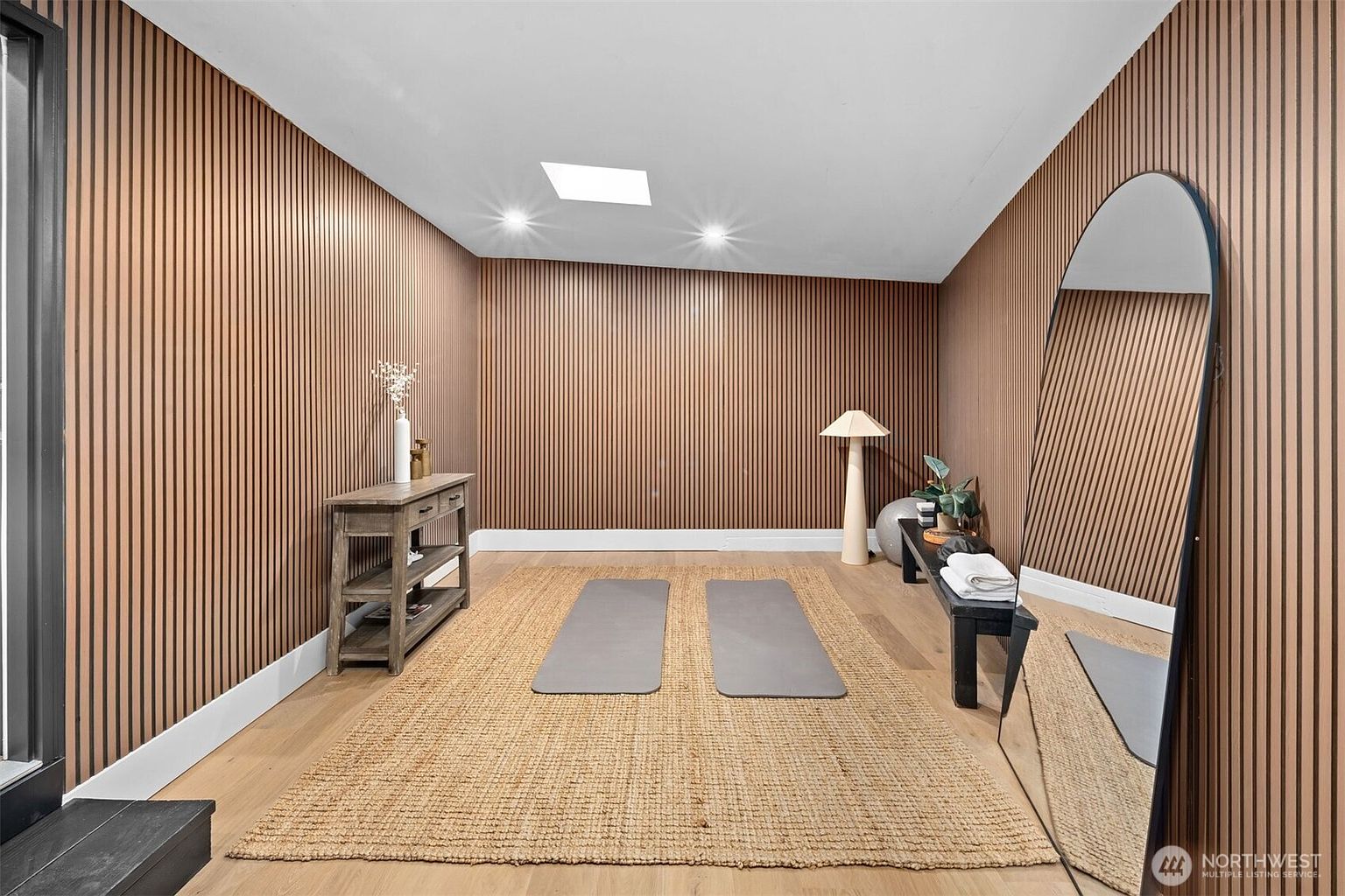 This serene home gym features warm, vertical wood-slat wall paneling that creates a textured, modern aesthetic. The space is centered around a large natural fiber rug holding two yoga mats, with a rustic wooden console table on the left and a black bench with exercise equipment on the right. A large, arched floor mirror reflects the room, while a skylight and recessed lighting provide bright, even illumination throughout the area.
