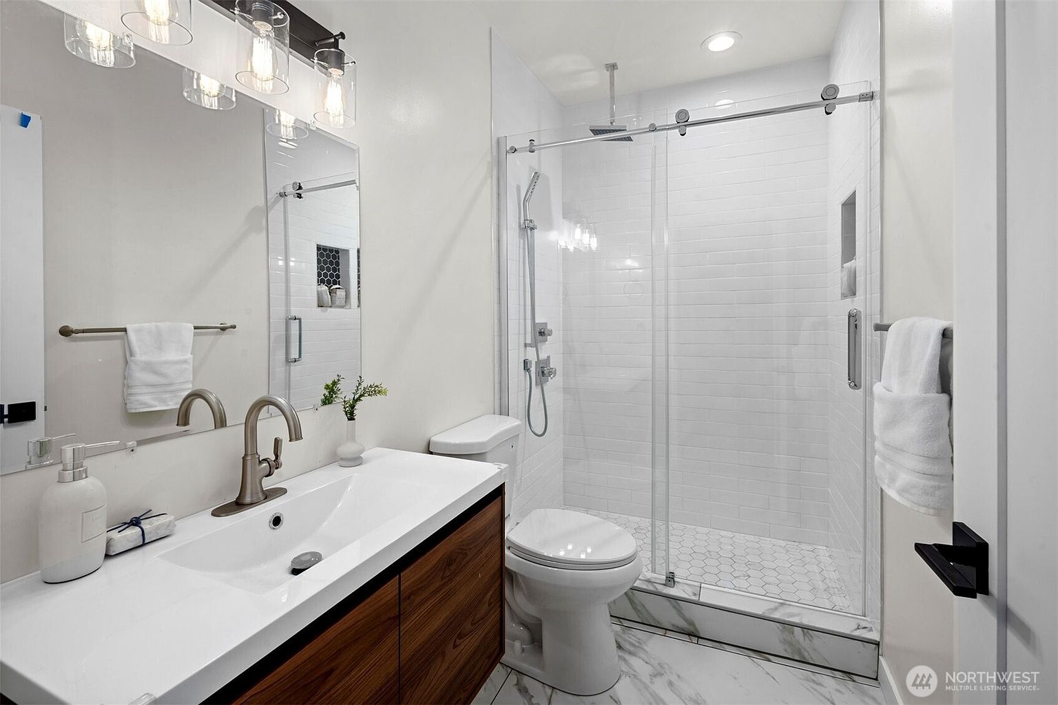 This modern bathroom features a floating vanity with a wood-toned cabinet and a white countertop, paired with a wall-mounted faucet and a large mirror. A glass-enclosed shower with white subway tile walls and a hexagonal tile floor sits adjacent to the toilet. The space is bright and clean, utilizing a neutral color palette and contemporary fixtures to create a functional and stylish aesthetic.
