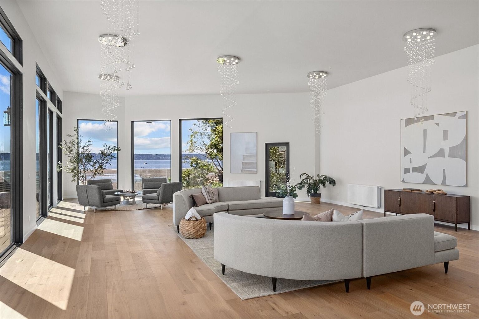 This spacious, modern living room features light wood flooring and a neutral color palette, anchored by a large, curved sectional sofa and two gray armchairs. The room is illuminated by three distinctive, cascading crystal pendant lights and benefits from abundant natural light provided by large windows that offer a scenic view of the water. A dark wood console table sits against the wall, complemented by minimalist artwork, creating an airy and sophisticated atmosphere.