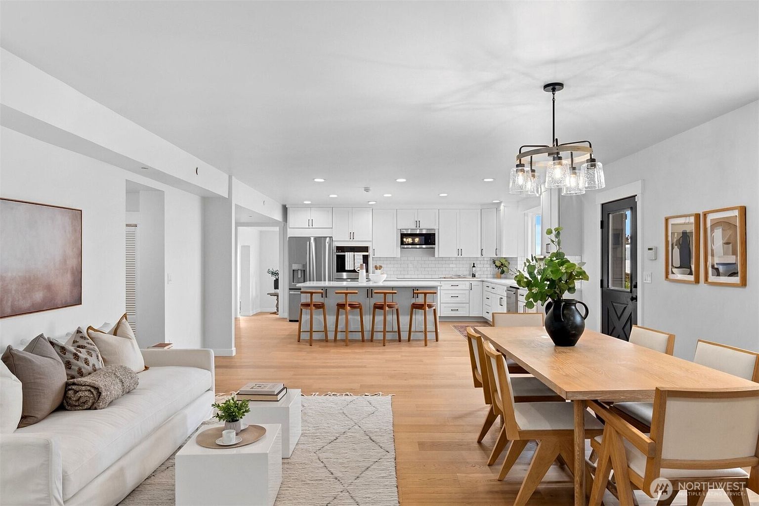 This open-concept living space features a bright, modern aesthetic with light wood flooring and a neutral color palette. The area seamlessly transitions from a cozy living room with a plush sofa to a dining space and a spacious kitchen equipped with a large island and white cabinetry. The perspective is captured from a wide-angle view, emphasizing the airy, interconnected layout of the home.