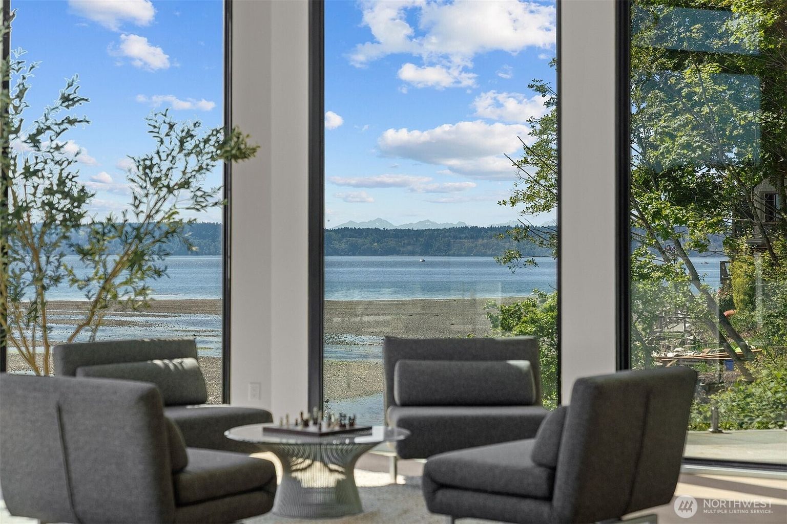 This living area features a sophisticated seating arrangement with four plush, dark gray armchairs centered around a modern, pedestal-base coffee table holding a chessboard. The space is defined by expansive floor-to-ceiling windows that frame a breathtaking, panoramic view of the water, shoreline, and distant mountains. The minimalist design emphasizes the connection between the interior living space and the natural landscape outside.