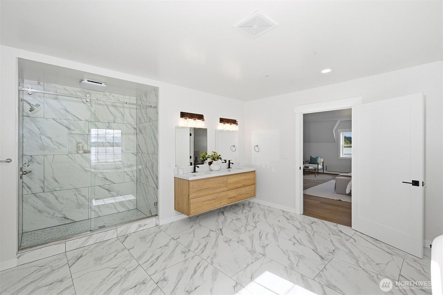 This bright, modern primary bathroom features a large walk-in shower with glass doors and patterned stone tile walls. A floating light wood vanity with dual sinks and dark fixtures is centered against the wall, complemented by two rectangular mirrors and wall-mounted lighting. The room is finished with matching patterned tile flooring and a doorway leading into an adjacent bedroom space.
