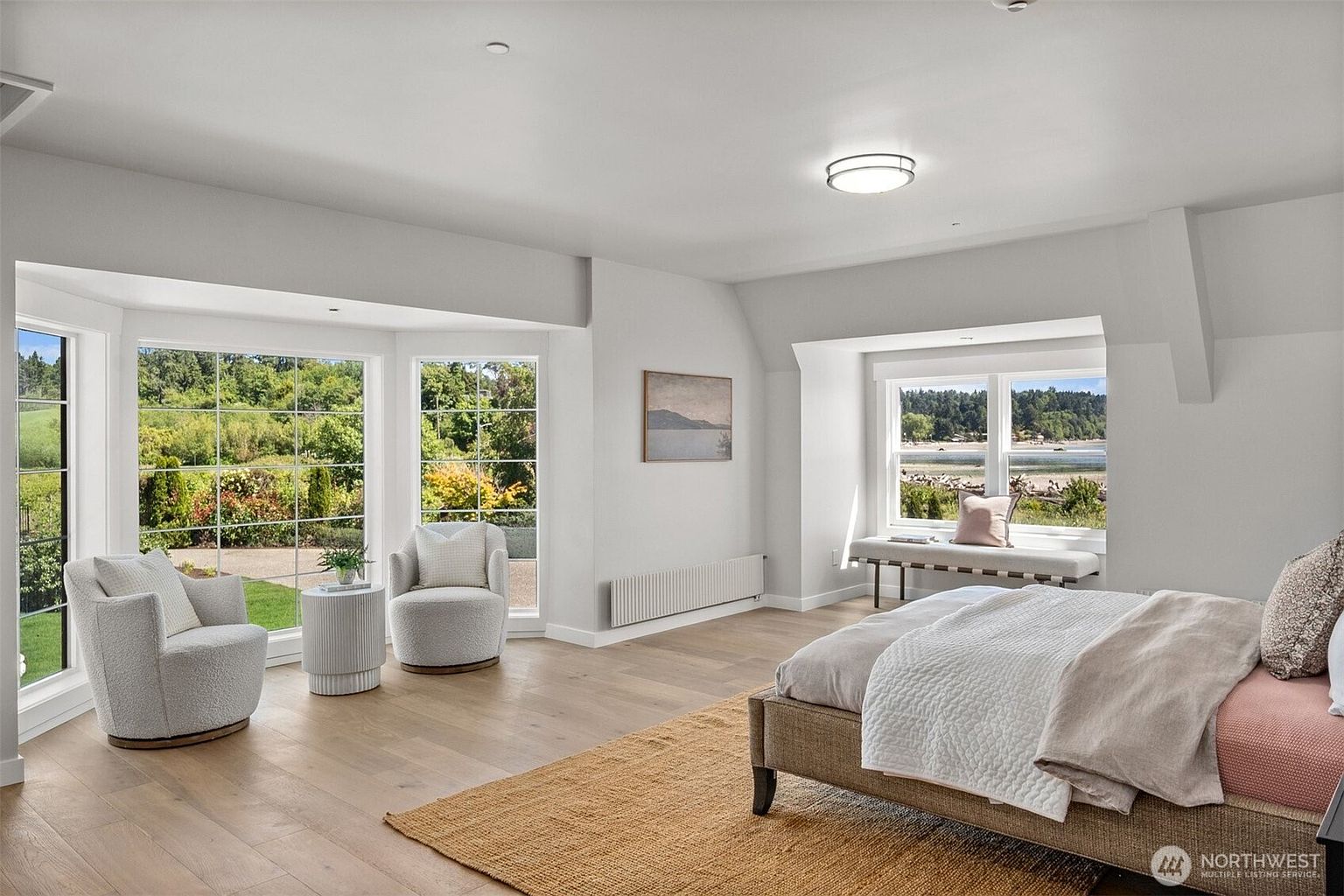 This spacious primary bedroom features light wood flooring, neutral walls, and a large bed positioned to face a window seat overlooking the water. To the left, a bay window area creates a cozy seating nook with two armchairs and a small side table, offering expansive views of the lush green landscape. The room is bright and airy, with a minimalist aesthetic and a textured rug anchoring the bed area.
