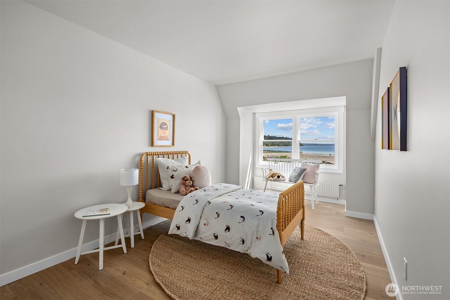 This bright and airy bedroom features light wood flooring, neutral walls, and a charming spindle-style bed frame. A large window provides a scenic view of the beach and water, while a round jute rug and a small side table add texture to the space. The room is decorated with playful artwork and soft textiles, creating a peaceful and inviting atmosphere.