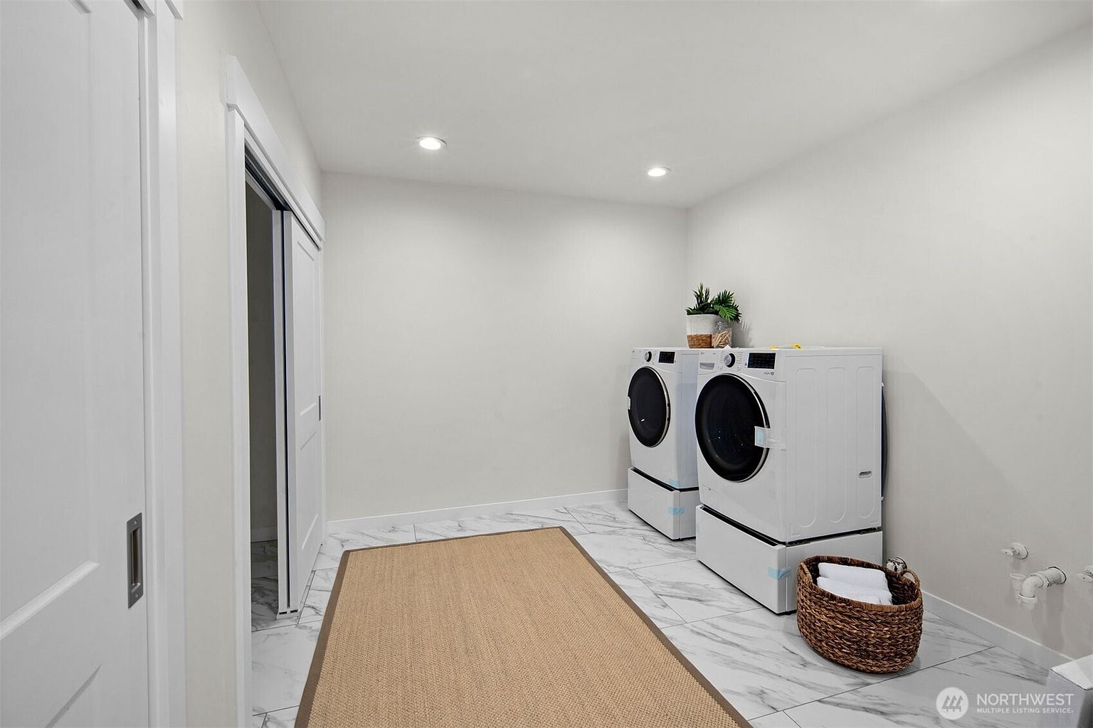 This clean and modern laundry room features white appliances, including a washer and dryer set, positioned against a plain white wall. The space is finished with light-colored, large-format patterned tile flooring and a neutral-toned area rug, creating a bright and functional atmosphere. A woven basket sits on the floor, and recessed lighting provides even illumination throughout the room.