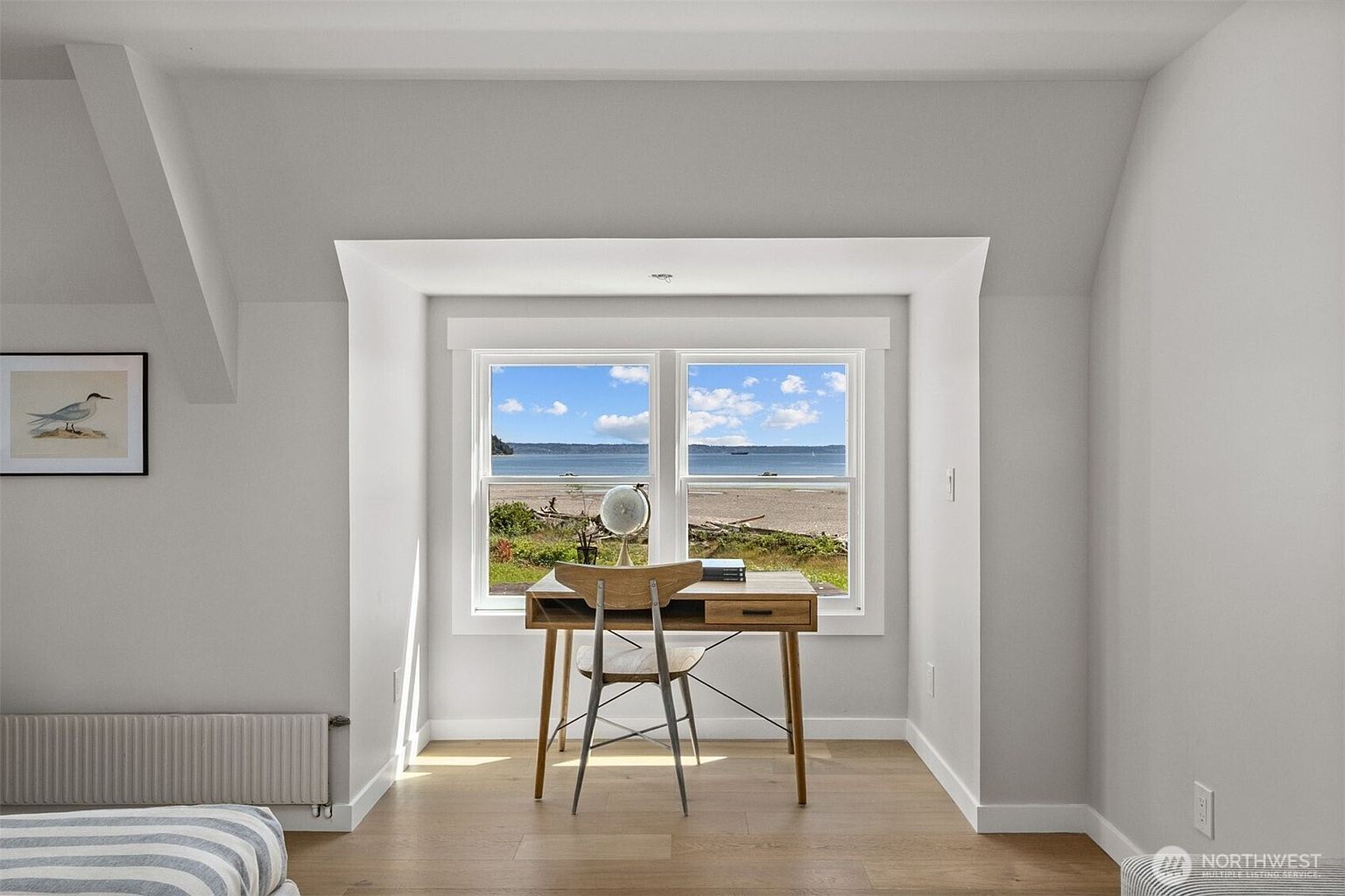 This bright and serene home office space is centered around a window nook that offers a picturesque view of the beach and ocean. The room features light-toned wood flooring, neutral walls, and a minimalist wooden desk paired with a modern chair. A framed bird print on the wall adds a touch of coastal charm to this tranquil, sunlit workspace.