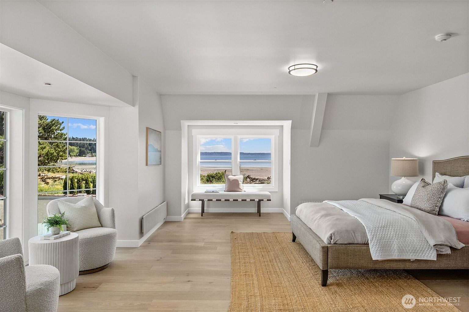 This bright and airy primary bedroom features light wood flooring, neutral walls, and a large bed with a textured headboard. A cozy seating area with two armchairs and a small side table sits near large windows, while a bench is positioned under a window nook that offers a scenic view of the beach and water. The room is illuminated by a modern flush-mount ceiling light, creating a serene and inviting atmosphere.