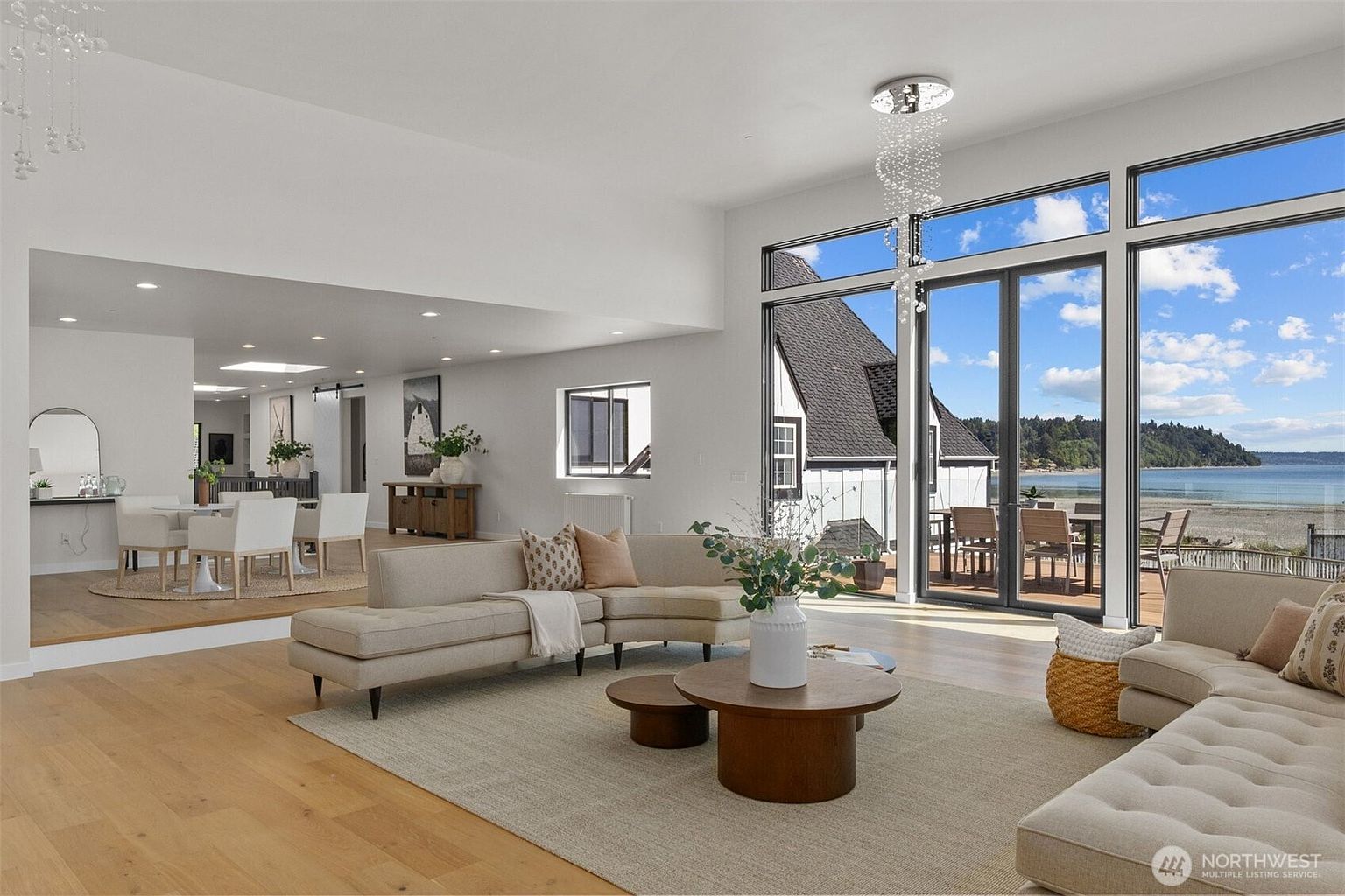 This bright, open-concept living area features a curved sectional sofa arranged on a large area rug, centered around a round wooden coffee table. The space is defined by light wood flooring and expansive glass doors that provide a seamless transition to an outdoor deck with panoramic water views. A dining area is visible on a slightly elevated platform in the background, creating a cohesive and airy atmosphere throughout the home.
