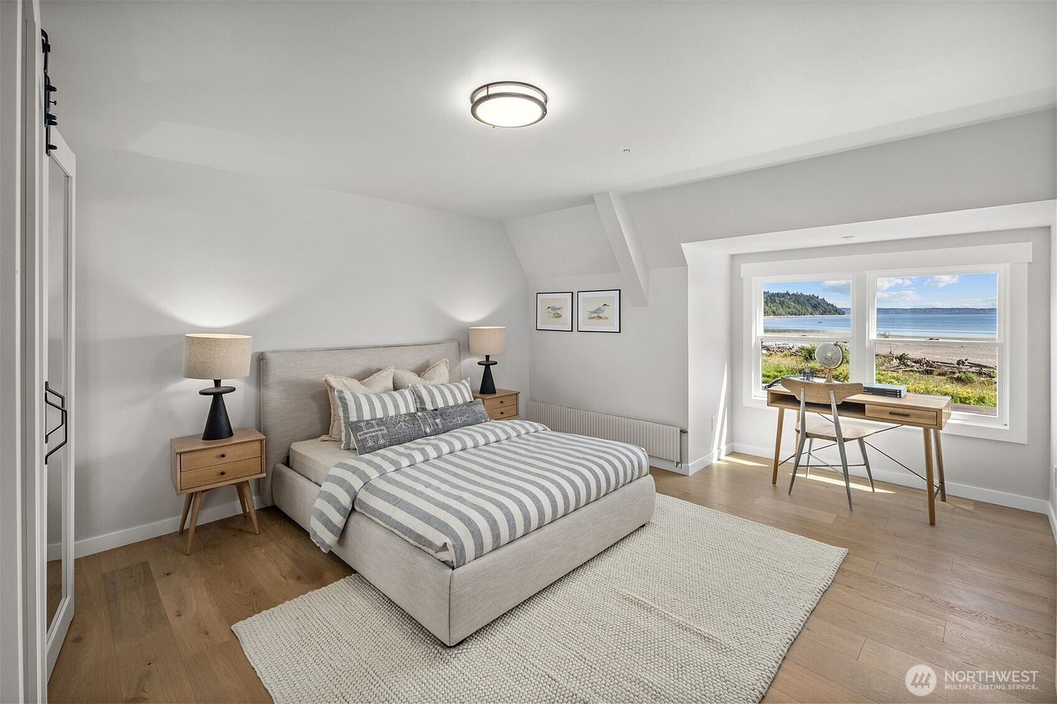 This bright and airy bedroom features a neutral color palette with light wood flooring and a textured area rug. A central bed with striped bedding is flanked by two matching nightstands and lamps, while a desk positioned near the large windows offers a scenic view of the beach and water. The room is finished with minimalist wall art and a clean, modern aesthetic that emphasizes natural light.