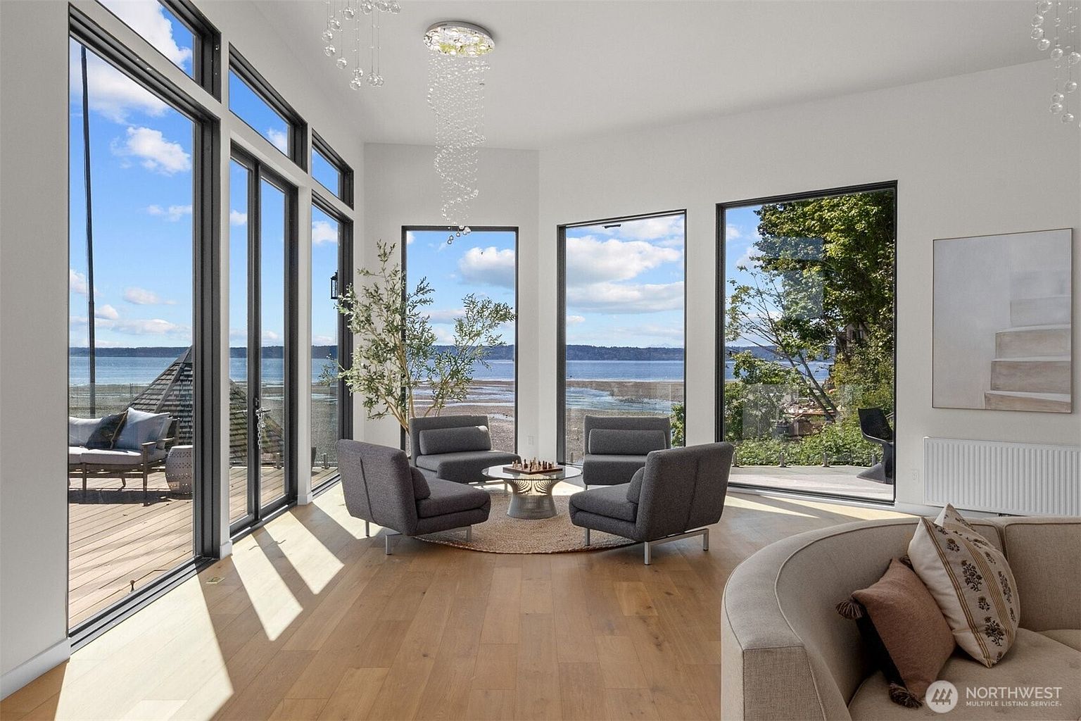 This bright and airy living room features light wood flooring and a sophisticated seating arrangement centered around a small chess table. Large windows provide expansive, unobstructed views of the water and shoreline, creating a seamless connection between the interior and the natural landscape. A modern, multi-tiered crystal chandelier hangs above the seating area, adding a touch of elegance to the minimalist, contemporary space.