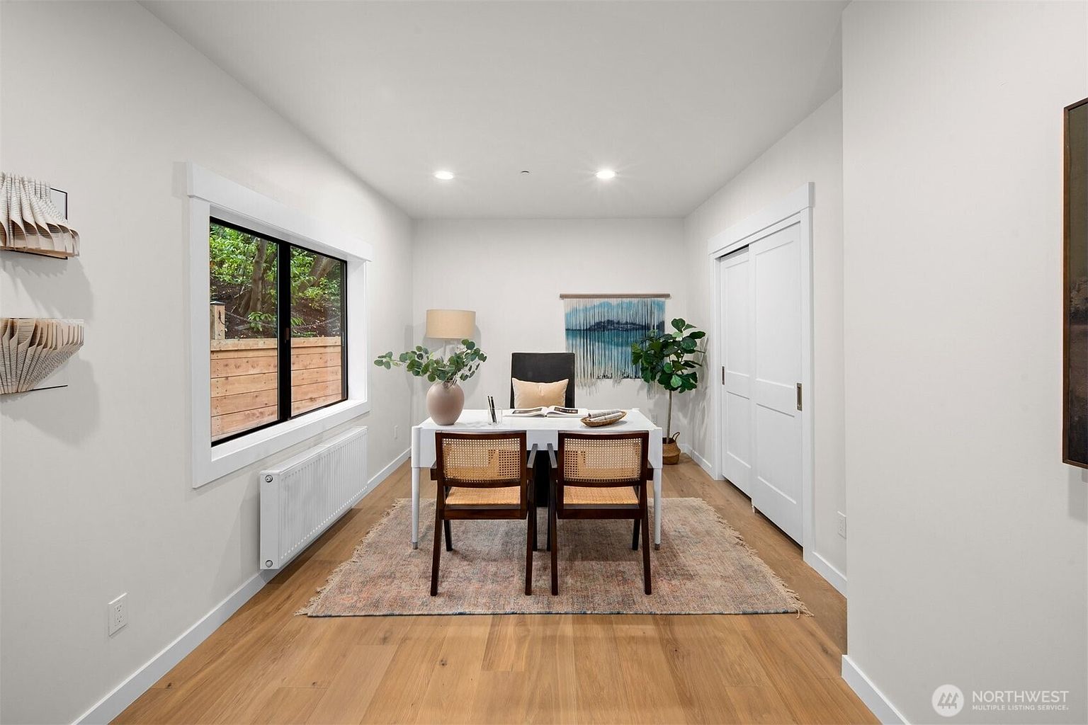 This bright and airy home office features light wood flooring, crisp white walls, and a large window that allows for ample natural light. The room is furnished with a sleek white desk, two woven-back chairs, and a decorative wall hanging, creating a clean and professional workspace. A closet with white double doors provides convenient storage, while the overall minimalist aesthetic offers a calm and productive atmosphere.