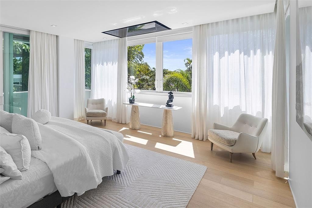 This bright and airy primary bedroom features a minimalist, contemporary design with floor-to-ceiling sheer curtains that flood the space with natural light. The room is anchored by a plush white bed and a sleek console table with sculptural accents, all set against light hardwood flooring and a textured area rug. The perspective captures a serene, high-end atmosphere with a clear view of lush tropical greenery outside the windows.