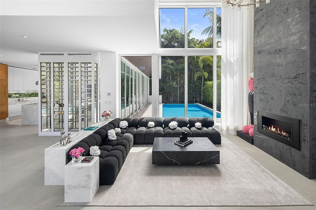 This sophisticated, modern living room features a large, dark-toned modular sofa arranged around a sleek black marble coffee table, set against a striking floor-to-ceiling textured gray fireplace wall. The space is bathed in natural light from expansive windows that offer a clear view of an outdoor swimming pool and lush greenery. A glass-enclosed wine display adds a touch of luxury, creating an open-concept layout that feels both spacious and high-end.