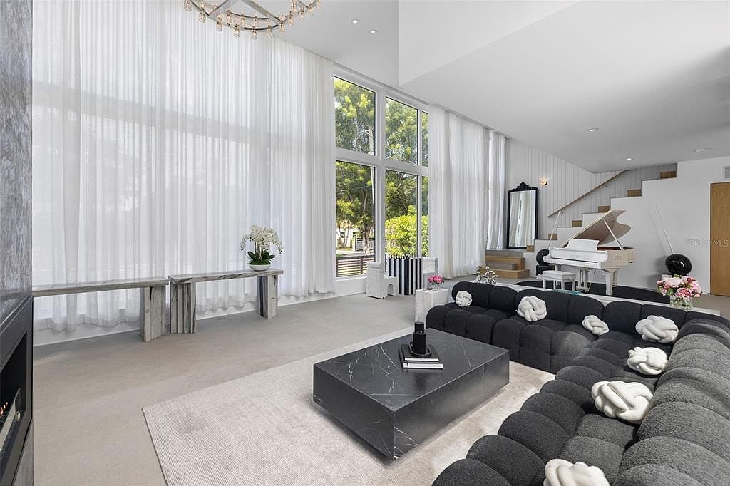 This expansive, high-ceilinged living room features a modern, minimalist aesthetic with a large, dark grey modular sofa and a striking black marble coffee table. The space is flooded with natural light from floor-to-ceiling windows draped in sheer white curtains, while a white grand piano adds a touch of elegance near the staircase. The overall impression is one of sophisticated, airy luxury with a clean, monochromatic color palette.