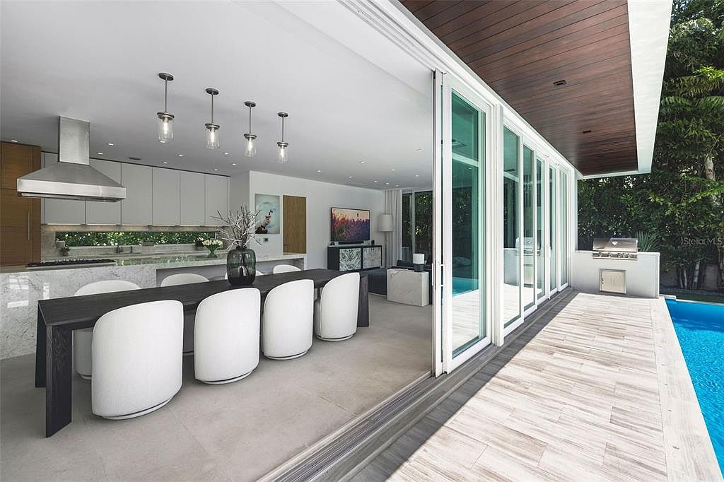 This modern, open-concept kitchen and dining area seamlessly transitions to an outdoor pool deck through expansive floor-to-ceiling sliding glass doors. The space features a sleek, dark wood dining table paired with minimalist white chairs, complemented by a contemporary kitchen island and high-end stainless steel appliances. The warm wood-paneled ceiling extending from the interior to the exterior patio creates a cohesive, luxurious indoor-outdoor living experience.