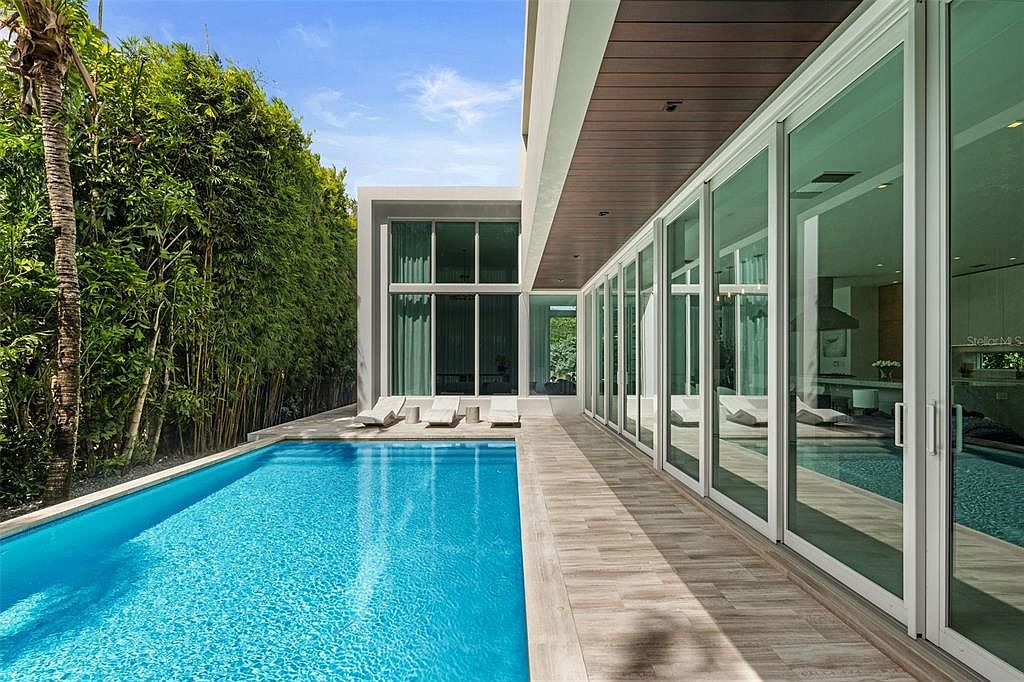 This modern, luxury residence features a sleek, rectangular swimming pool set against a backdrop of lush, tall bamboo privacy hedges. A wide, light-toned tiled patio runs alongside the house, which is characterized by floor-to-ceiling glass sliding doors that seamlessly blend indoor and outdoor living spaces. The architectural design is contemporary and minimalist, emphasizing clean lines and a sophisticated, resort-like atmosphere.