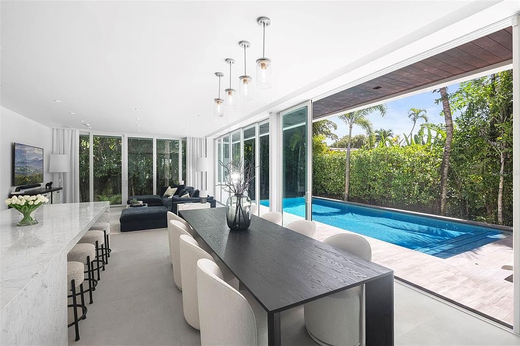 This modern, open-concept dining area features a sleek black table paired with light-colored upholstered chairs, seamlessly transitioning to an outdoor pool area through floor-to-ceiling glass sliding doors. The space is illuminated by contemporary pendant lighting and includes a marble-topped kitchen island with bar seating, creating a sophisticated and airy atmosphere. The view highlights the integration of indoor and outdoor living, perfect for entertaining in a luxury tropical setting.