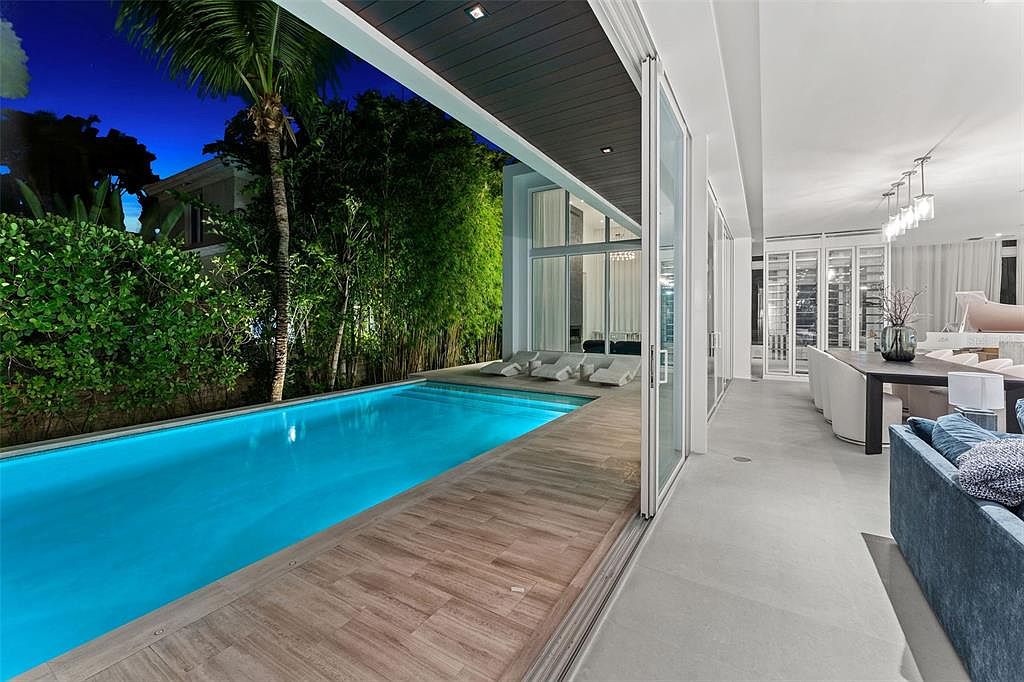 This stunning contemporary outdoor space features a sleek, rectangular swimming pool bordered by a warm-toned wood deck and a covered patio area. Large sliding glass doors seamlessly connect the indoor living and dining areas to the pool deck, creating an expansive open-concept transition. The scene is captured at twilight, highlighting the vibrant blue pool water and the lush, illuminated bamboo landscaping that provides a private, resort-like atmosphere.