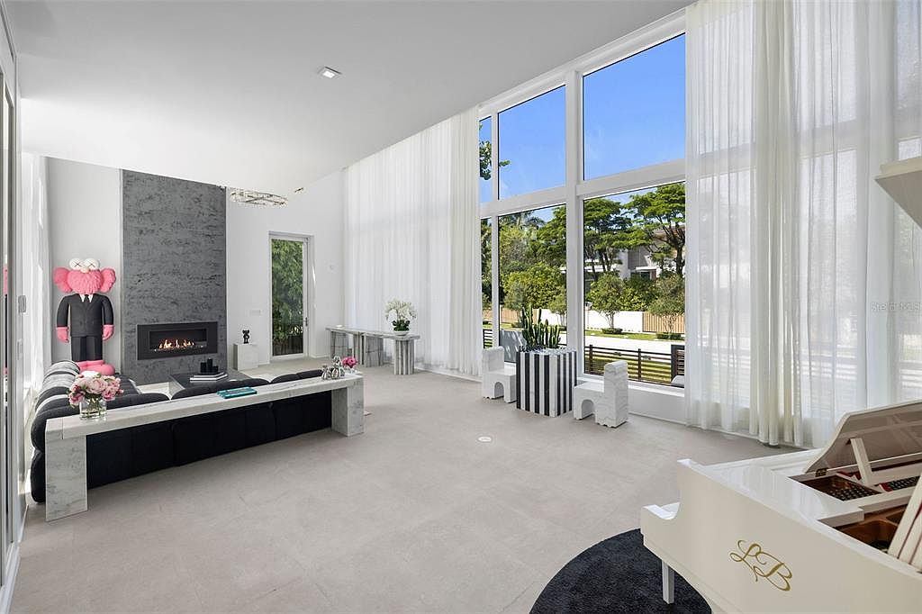 This expansive, high-ceilinged living room features a modern aesthetic with a striking gray stone fireplace, a large black sofa, and a white grand piano. Floor-to-ceiling windows draped in sheer curtains flood the space with natural light, offering a view of the lush exterior greenery. The room is accented with unique art pieces, including a large pink KAWS figure, creating a sophisticated and contemporary atmosphere.