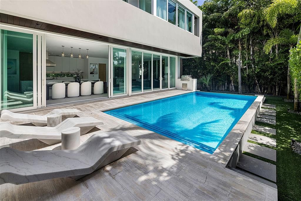 This modern, high-end residence features a seamless indoor-outdoor transition with large sliding glass doors opening onto a sleek stone patio. The centerpiece is a vibrant blue rectangular swimming pool, complemented by contemporary stone lounge chairs and a lush, private backyard setting. The architectural style is minimalist and sophisticated, emphasizing clean lines and luxury living.