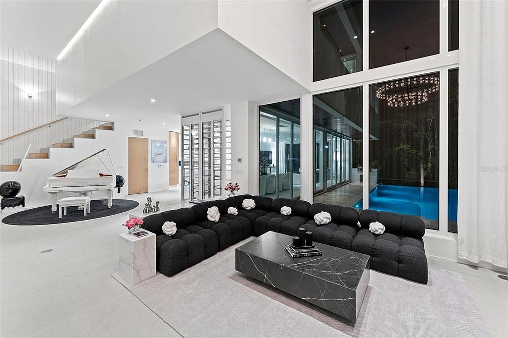This expansive, ultra-modern living room features a double-height ceiling and floor-to-ceiling windows that offer a seamless view of an illuminated outdoor pool. The space is anchored by a large, black modular sectional sofa and a striking black marble coffee table, complemented by a white grand piano positioned near a floating staircase. The minimalist, high-contrast aesthetic creates a sophisticated and luxurious atmosphere perfect for high-end entertaining.