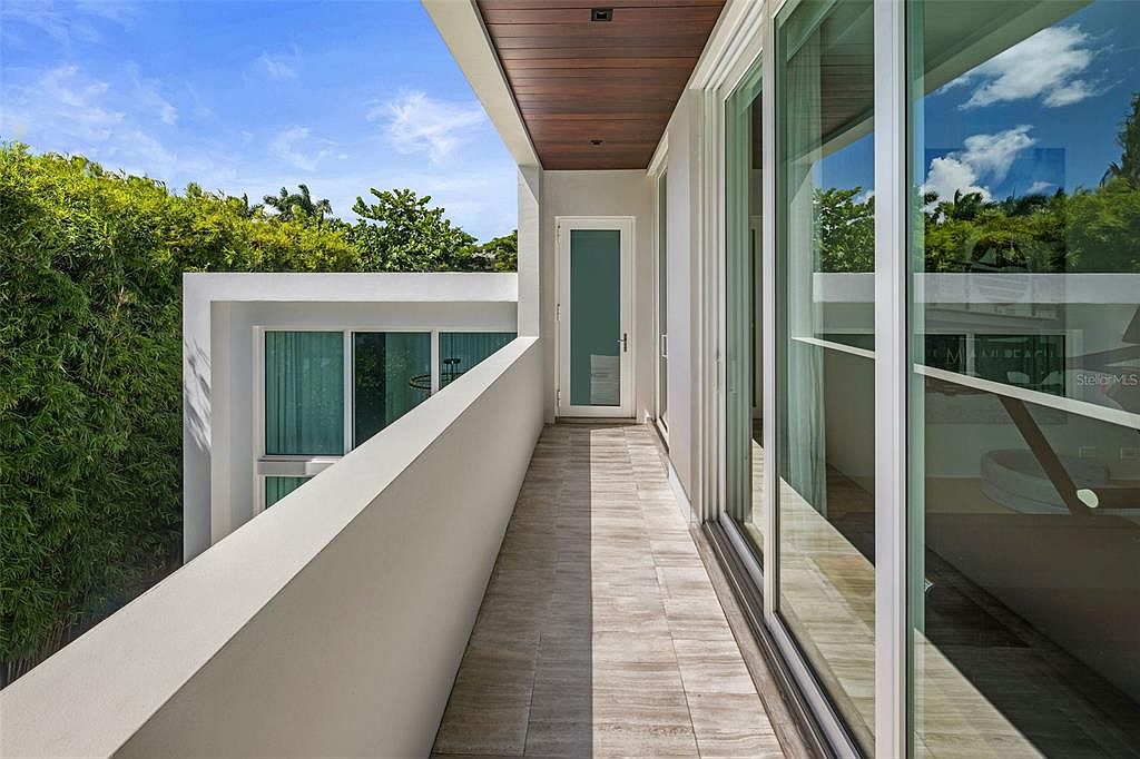 This modern balcony features sleek, light-toned tile flooring and a minimalist white railing that overlooks lush green foliage. A dark wood-paneled ceiling adds warmth to the space, while large sliding glass doors provide seamless indoor-outdoor connectivity. The perspective captures the clean lines and contemporary architectural design of the home's exterior.