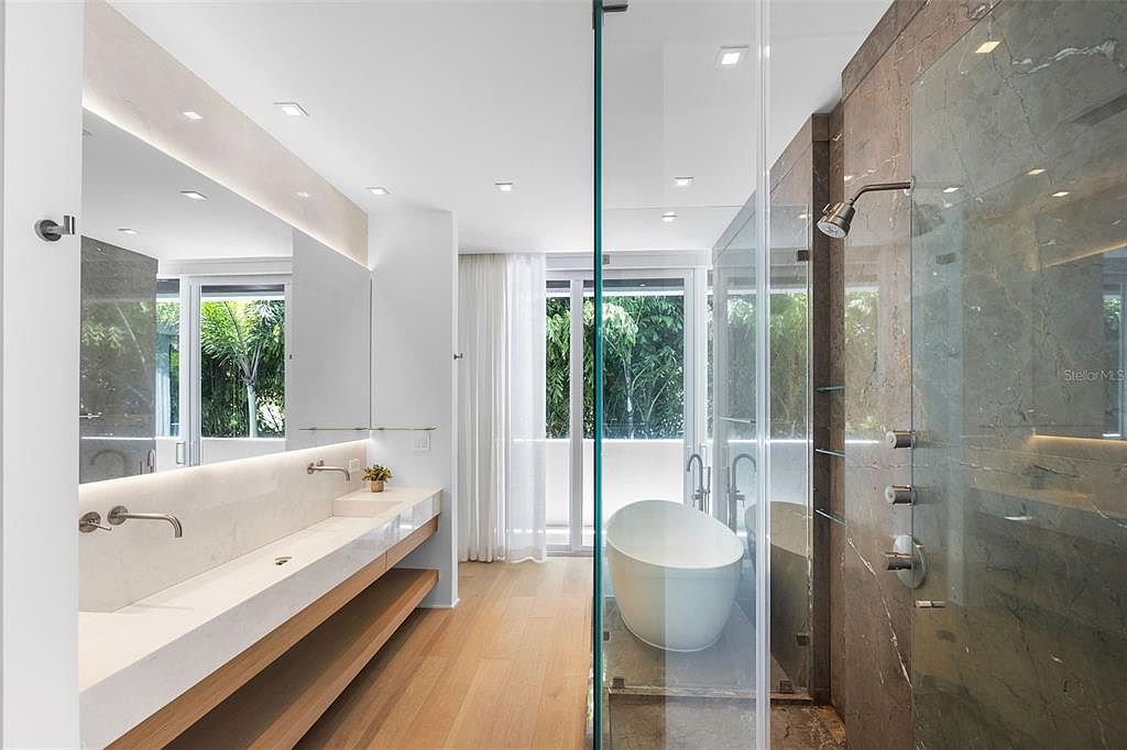 This luxurious primary bathroom features a sleek, modern design with a floating double vanity, light wood flooring, and a spacious walk-in shower enclosed by glass. A freestanding soaking tub sits elegantly near floor-to-ceiling windows that offer a view of lush greenery, creating a serene, spa-like atmosphere. The combination of warm wood tones and cool stone surfaces provides a sophisticated and high-end aesthetic.