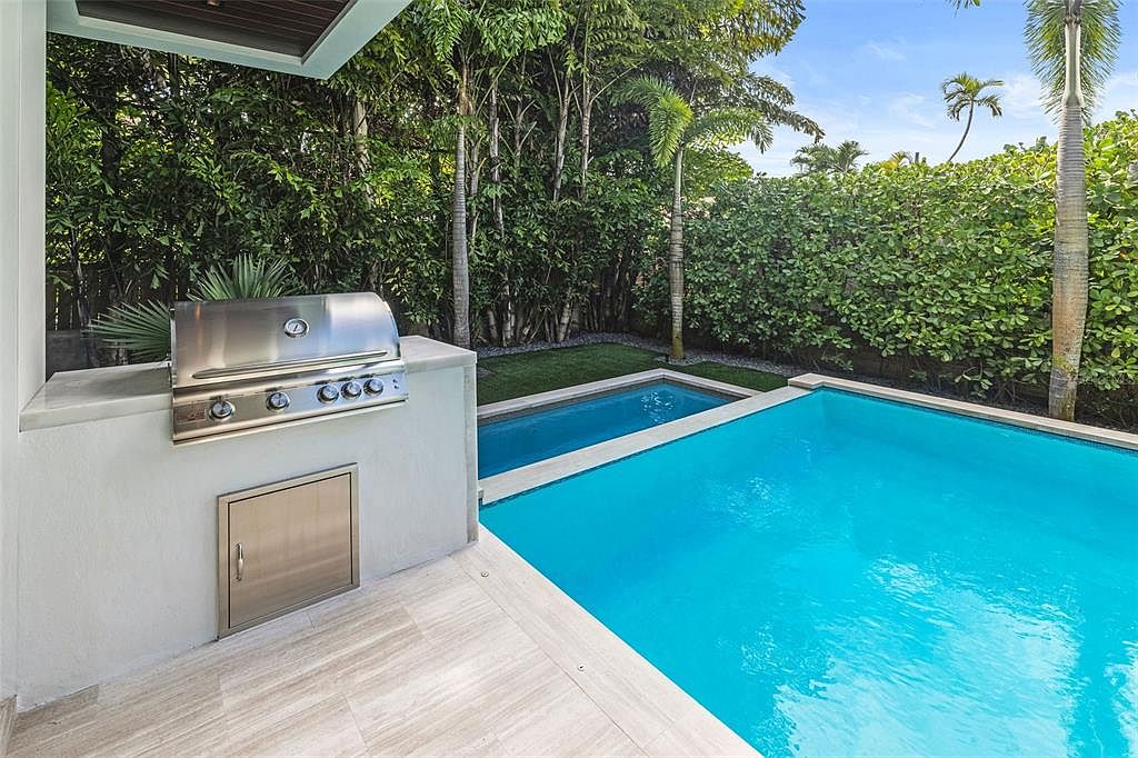 This image captures a modern outdoor living space featuring a sleek, built-in stainless steel grill set into a light-colored stucco counter. Adjacent to the grill is a vibrant, crystal-clear swimming pool surrounded by light-toned stone decking and lush tropical landscaping. The perspective is from a covered patio area, looking out toward the pool and the dense green hedge that provides privacy for the backyard.
