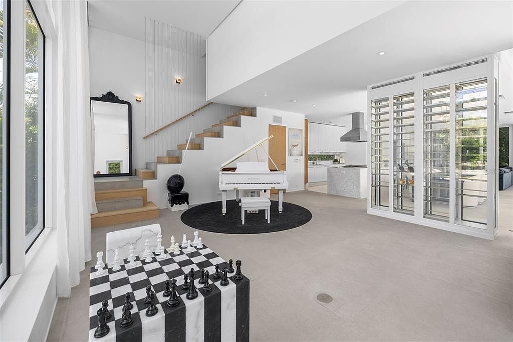 This expansive, modern open-concept living area features a striking white grand piano as its centerpiece, set upon a circular black rug. The space includes a unique oversized chess set in the foreground, a floating staircase with glass railings, and a sleek glass-enclosed wine display. The minimalist aesthetic is enhanced by high ceilings, neutral tones, and a seamless transition into a contemporary kitchen in the background.