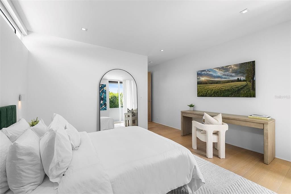 This bright and airy primary bedroom features a minimalist aesthetic with crisp white bedding, light wood flooring, and a large arched floor mirror. A sleek, wall-mounted television sits above a long, modern wooden console table paired with a textured white accent chair. The room is bathed in natural light, creating a serene and sophisticated atmosphere.