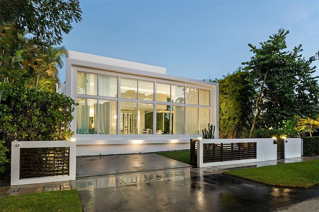 This striking modern residence features a minimalist white facade dominated by expansive floor-to-ceiling glass windows that offer a clear view into the illuminated interior. The property is framed by a sleek, contemporary wall with horizontal slat detailing and lush tropical landscaping, creating a sophisticated curb appeal. The wet pavement in the foreground suggests a recent rain, adding a reflective, cinematic quality to the twilight setting.