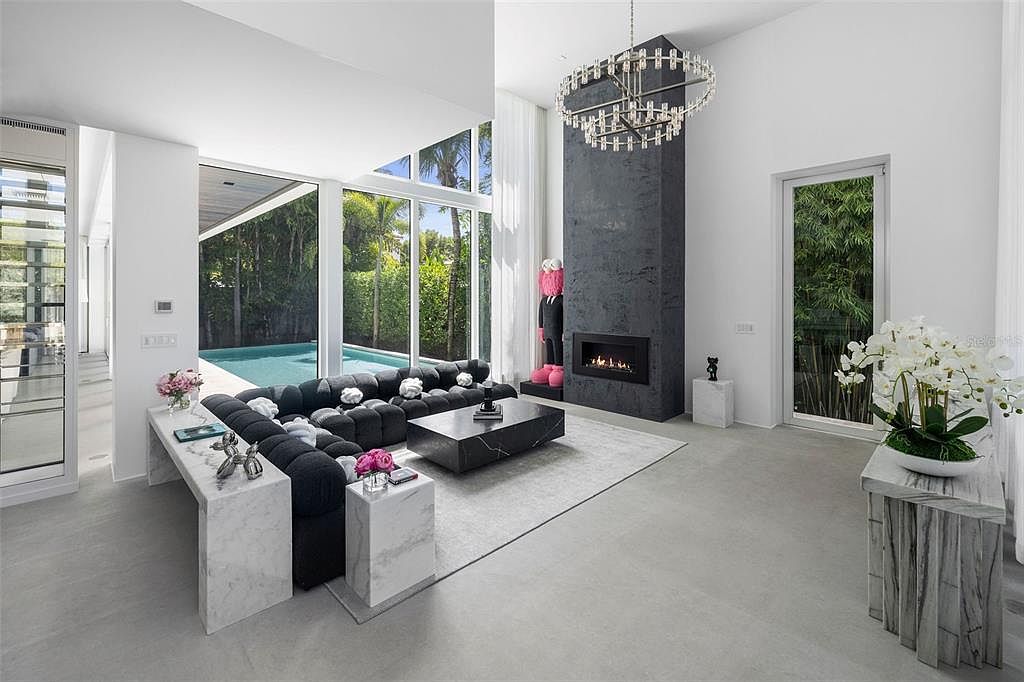 This sophisticated, high-ceiling living room features a striking black stone fireplace as its focal point, complemented by a plush, dark-toned modular sofa and a sleek marble coffee table. Large floor-to-ceiling windows provide an expansive view of the outdoor pool and lush greenery, creating a seamless indoor-outdoor living experience. The minimalist aesthetic is enhanced by elegant marble side tables, a contemporary chandelier, and a vibrant art piece, exuding a luxurious and modern atmosphere.