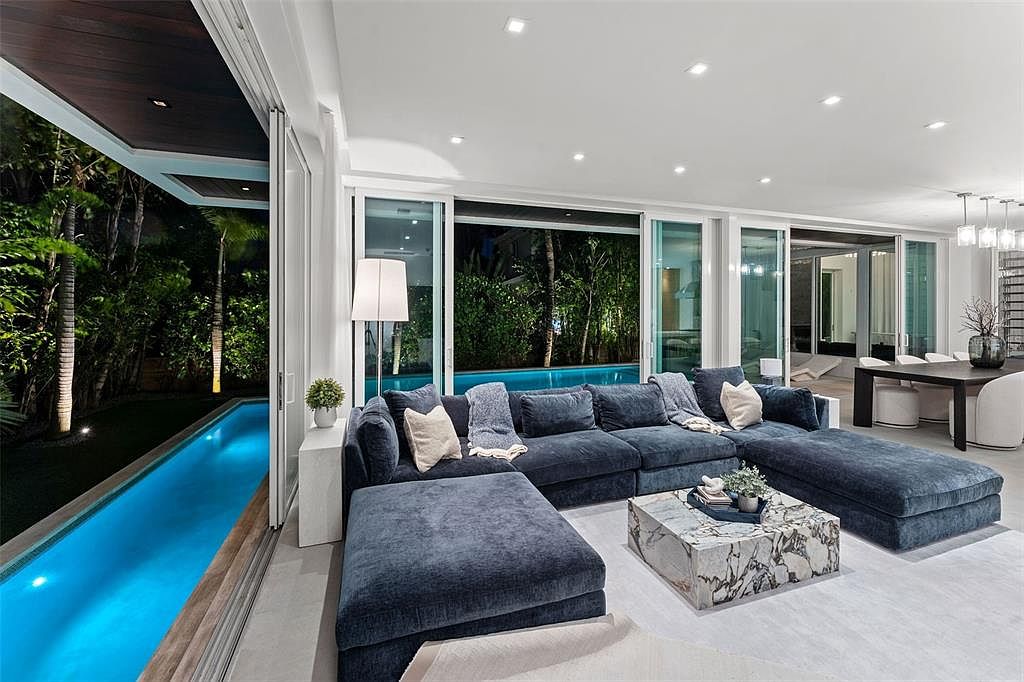 This sophisticated, open-concept living room features a large, plush navy blue sectional sofa and a striking marble coffee table, creating a luxurious and inviting atmosphere. The space is seamlessly connected to an outdoor lap pool through expansive floor-to-ceiling sliding glass doors, blurring the lines between indoor and outdoor living. The modern design is complemented by recessed lighting and a minimalist aesthetic, offering a serene view of the illuminated pool and lush landscaping at night.
