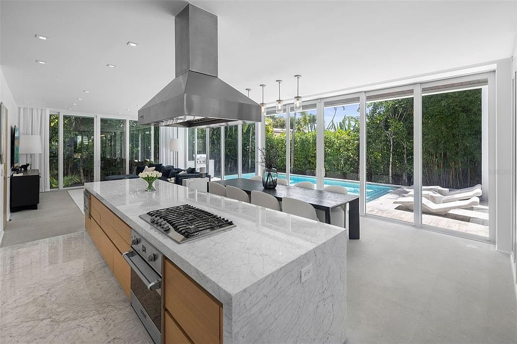 This sleek, modern kitchen features a large waterfall-edge marble island with a built-in gas cooktop and stainless steel range hood. The space flows seamlessly into a dining area and opens up to a bright, sunlit patio with a swimming pool through floor-to-ceiling glass sliding doors. The minimalist design, light wood cabinetry, and polished stone flooring create an airy, luxurious atmosphere perfect for indoor-outdoor living.