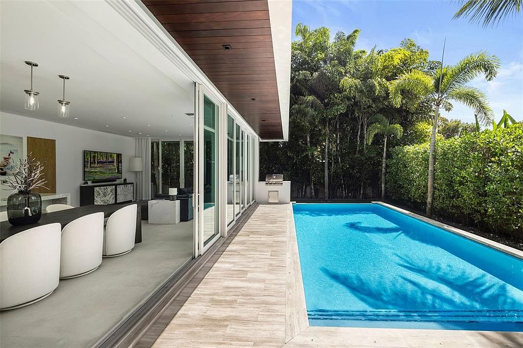 This image captures a seamless transition between a modern indoor dining area and a luxurious outdoor pool deck. The space features floor-to-ceiling sliding glass doors that open onto a tiled patio, leading directly to a vibrant blue swimming pool surrounded by lush tropical greenery. The architectural design emphasizes an open-concept flow, blending contemporary interior aesthetics with a serene, resort-style outdoor living environment.