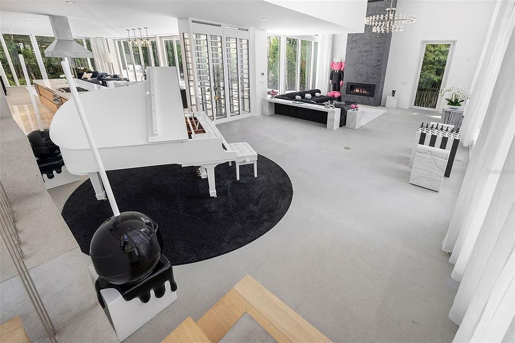 This expansive, ultra-modern living space features a high-contrast monochromatic design with a striking white grand piano as a focal point on a circular black rug. The room is characterized by soaring ceilings, floor-to-ceiling windows, and a sleek fireplace with a dark stone surround, creating a sophisticated and luxurious atmosphere. The open-concept layout seamlessly integrates a contemporary seating area and a dining space, emphasizing a minimalist and high-end aesthetic.