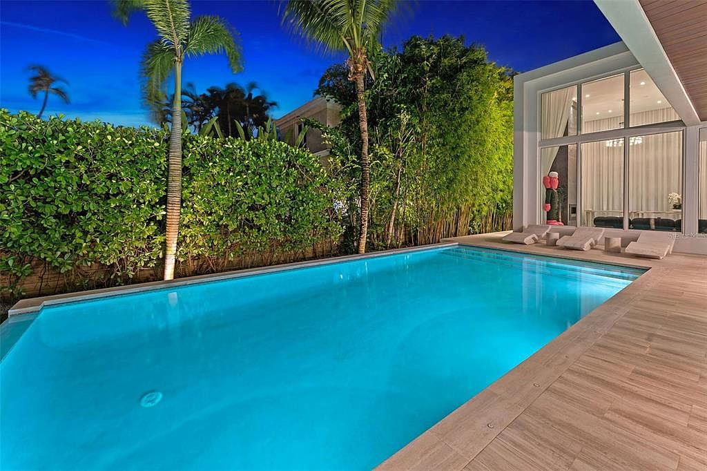 This stunning evening shot captures a modern, illuminated swimming pool set against a lush, private backyard landscape. The scene features sleek wooden decking, contemporary lounge chairs, and floor-to-ceiling glass walls that seamlessly connect the outdoor space to the home's interior. The vibrant blue pool lighting and the twilight sky create a sophisticated, resort-like atmosphere perfect for luxury living.
