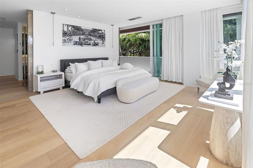 This bright and airy primary bedroom features a minimalist, contemporary design with a neutral color palette and light hardwood flooring. The room is anchored by a large bed with crisp white linens, a sleek bench, and a textured area rug, all illuminated by natural light streaming through floor-to-ceiling windows. A stylish console table with floral decor and a modern art piece above the bed add sophisticated touches to this serene, high-end living space.