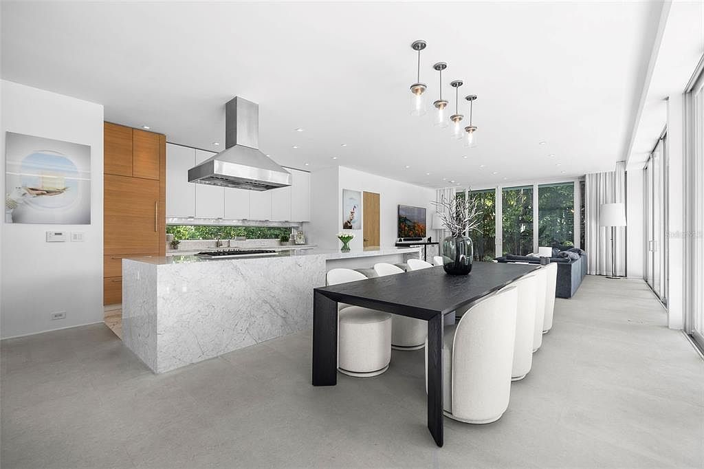 This modern, open-concept kitchen and dining area features a striking marble waterfall island and sleek white cabinetry paired with warm wood accents. A long, dark wood dining table sits in the foreground, surrounded by plush white chairs, while floor-to-ceiling glass doors provide a seamless transition to the outdoors. The space is illuminated by contemporary pendant lighting and recessed ceiling lights, creating a bright, sophisticated, and airy atmosphere.