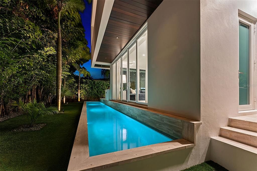 This image captures a sleek, modern lap pool illuminated at night, running parallel to a contemporary home with large glass sliding doors. The architectural design features a warm wood-paneled overhang and clean, minimalist lines, creating a sophisticated outdoor living space. The perspective highlights the seamless transition between the interior living area and the tranquil, glowing pool, set against a backdrop of lush, manicured landscaping.