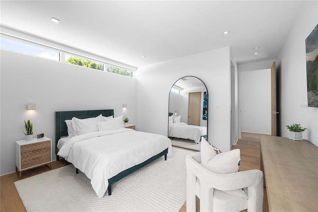 This bright and airy primary bedroom features a modern aesthetic with a dark green upholstered bed frame, crisp white bedding, and light wood flooring. A large arched floor mirror reflects the room, while a textured white armchair and a minimalist wooden nightstand add layers of comfort and style. The space is illuminated by natural light from high clerestory windows, creating a serene and sophisticated atmosphere.