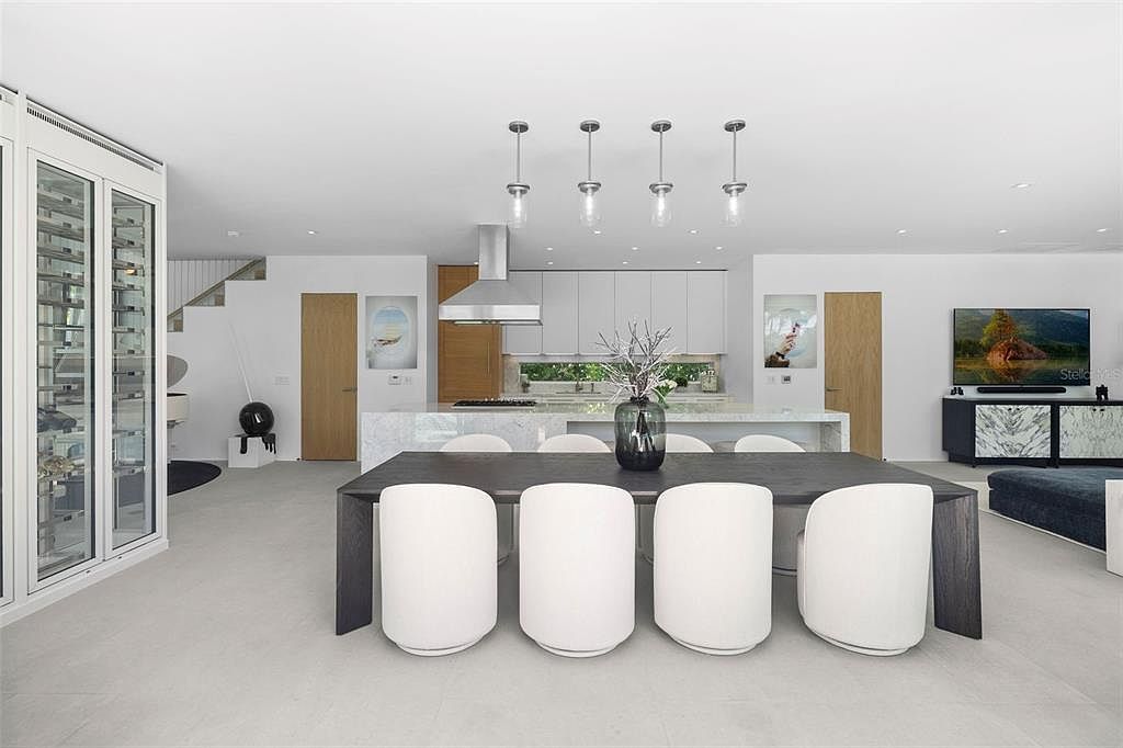This modern, open-concept dining area features a sleek, dark wood table paired with four minimalist white chairs, set against a backdrop of a contemporary kitchen with marble-topped island and white cabinetry. To the left, a glass-enclosed wine cellar adds a sophisticated touch, while a large television is mounted on the right wall. The space is illuminated by four elegant pendant lights, creating a bright, airy, and high-end atmosphere.