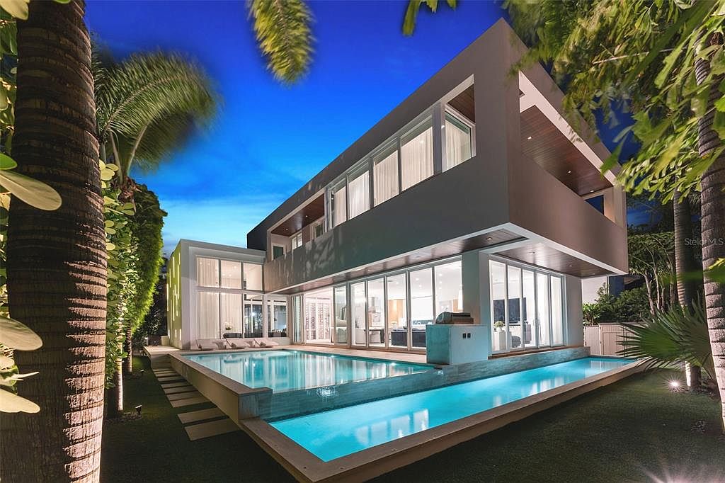 This stunning contemporary residence features a sleek, multi-level architectural design with expansive floor-to-ceiling glass walls that seamlessly blend indoor and outdoor living. The property is highlighted by a sophisticated, illuminated infinity-edge pool that wraps around the rear of the home, creating a luxurious resort-like atmosphere at twilight. Lush tropical landscaping and palm trees frame the structure, enhancing the modern aesthetic and providing a serene, high-end residential appeal.