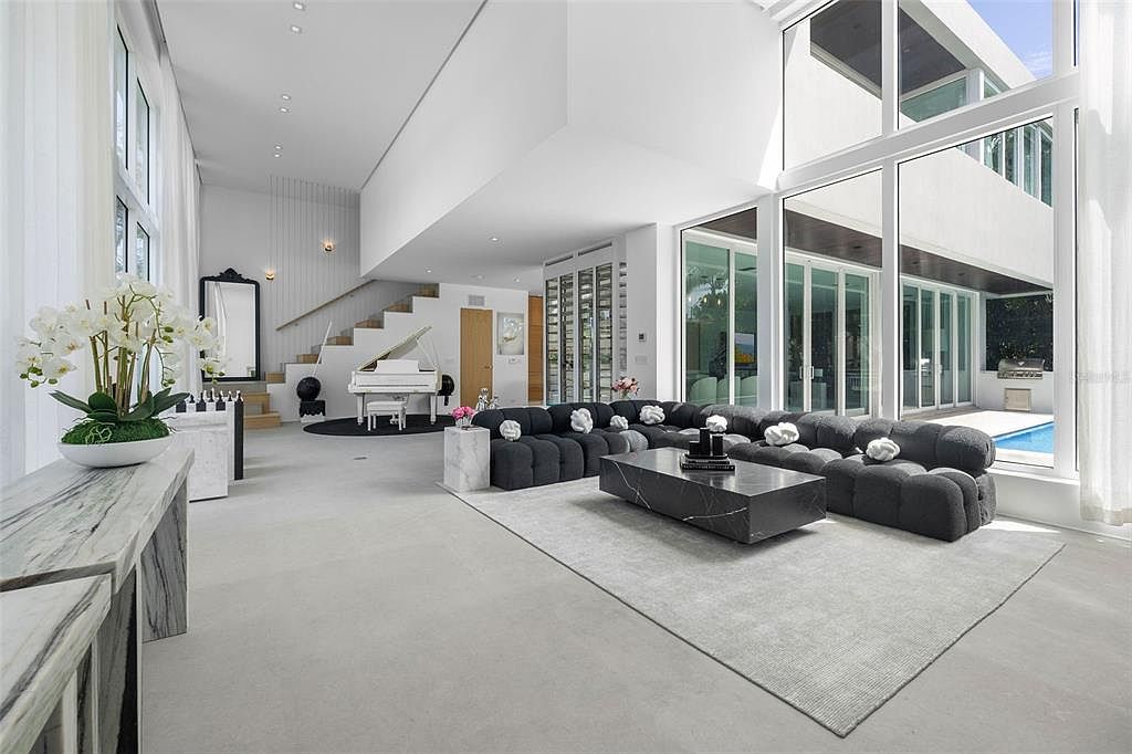 This expansive, double-height living room features a minimalist, modern aesthetic with a large, dark grey modular sofa as the centerpiece. The space is illuminated by floor-to-ceiling windows that offer a view of the outdoor pool area, while a white grand piano sits elegantly near the staircase. The clean lines, neutral color palette, and high-end finishes create a sophisticated and airy atmosphere.