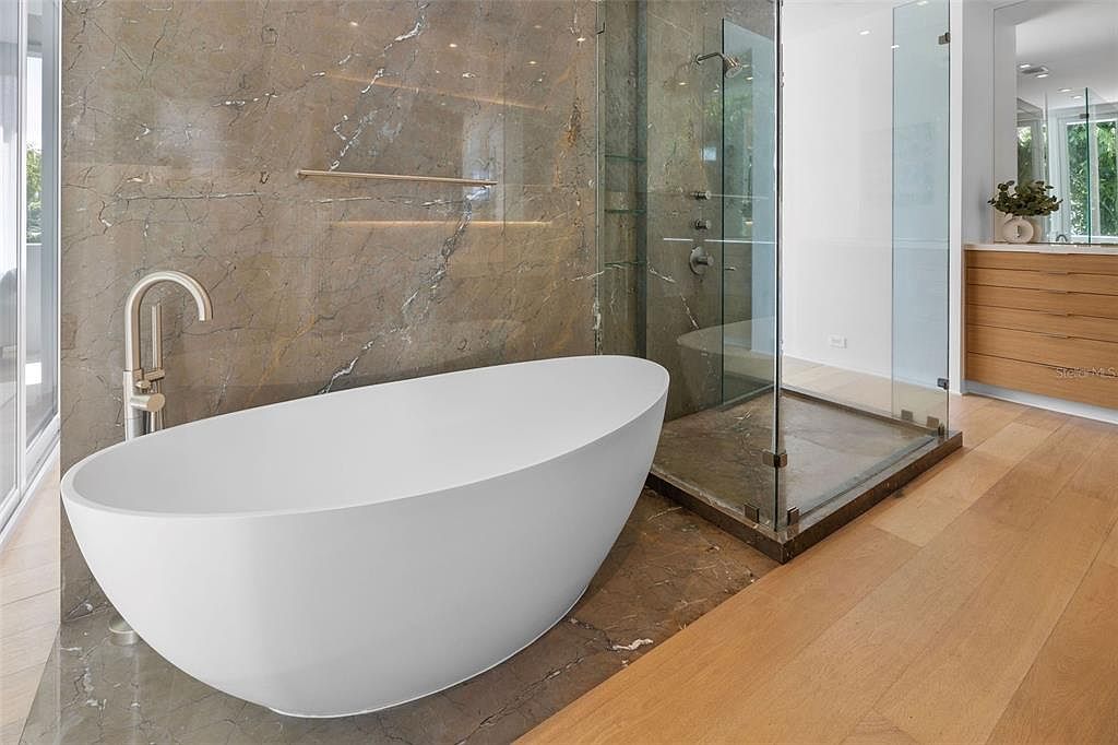 This luxurious primary bathroom features a sleek, white freestanding soaking tub set against a dramatic, large-format marble accent wall. Adjacent to the tub is a spacious walk-in glass shower with dark stone flooring, creating a sophisticated spa-like atmosphere. The room is finished with warm-toned hardwood flooring and a modern vanity, blending natural textures with contemporary design elements.