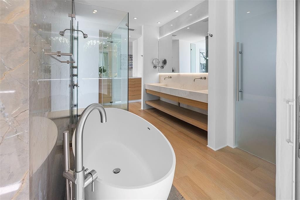This luxurious primary bathroom features a sleek, modern design with a freestanding white soaking tub as the focal point. The space includes a glass-enclosed walk-in shower, a double vanity with a floating wooden shelf, and light wood flooring that adds warmth to the clean, minimalist aesthetic. The perspective captures the open, airy layout, highlighting the high-end finishes and sophisticated lighting.