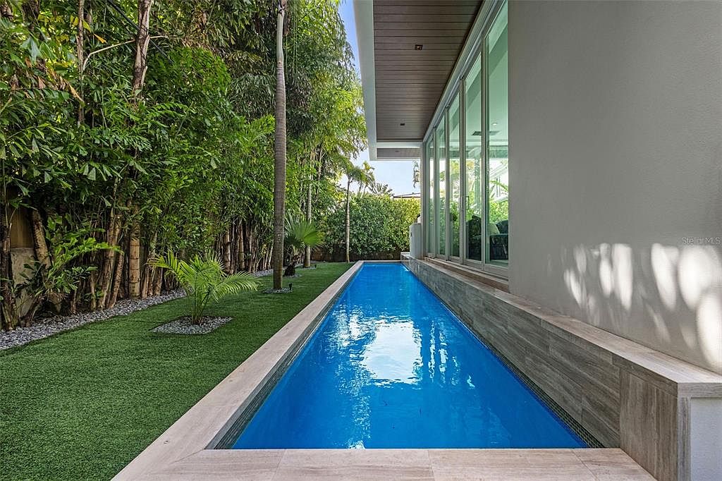 This image showcases a sleek, modern lap pool running parallel to the side of a contemporary home, featuring large floor-to-ceiling glass sliding doors. The pool is bordered by light-toned stone decking and a lush, manicured lawn lined with tall bamboo and palm trees. The perspective is a low-angle, eye-level shot that emphasizes the clean lines and tranquil, resort-like atmosphere of the outdoor space.