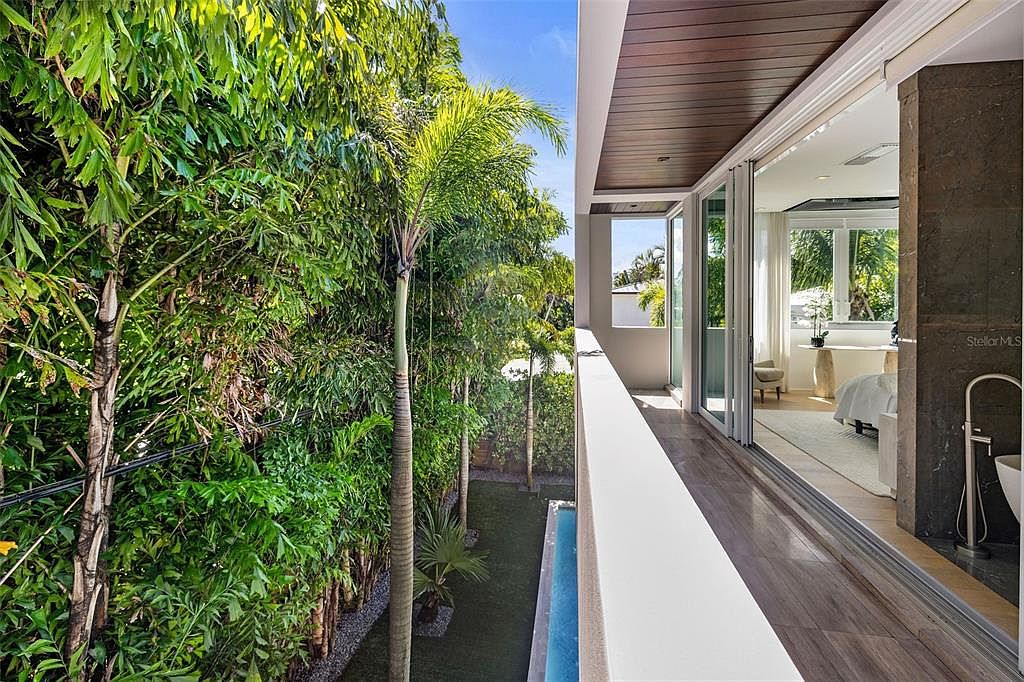 This image captures a sleek, modern balcony overlooking a lush, tropical backyard with a narrow lap pool. The balcony features dark wood-look flooring and a clean white railing, seamlessly connecting to an interior bedroom through large sliding glass doors. The perspective emphasizes the transition between the refined indoor living space and the vibrant, green outdoor environment.