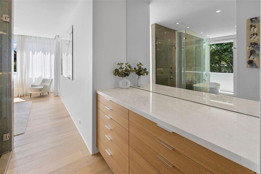 This modern primary bathroom features a sleek, light wood vanity with a white quartz countertop and a large, seamless mirror that reflects the adjacent shower area. The space is bright and airy, with light hardwood flooring extending into a hallway that leads to a sunlit seating area. The minimalist design and clean lines create a sophisticated, spa-like atmosphere.